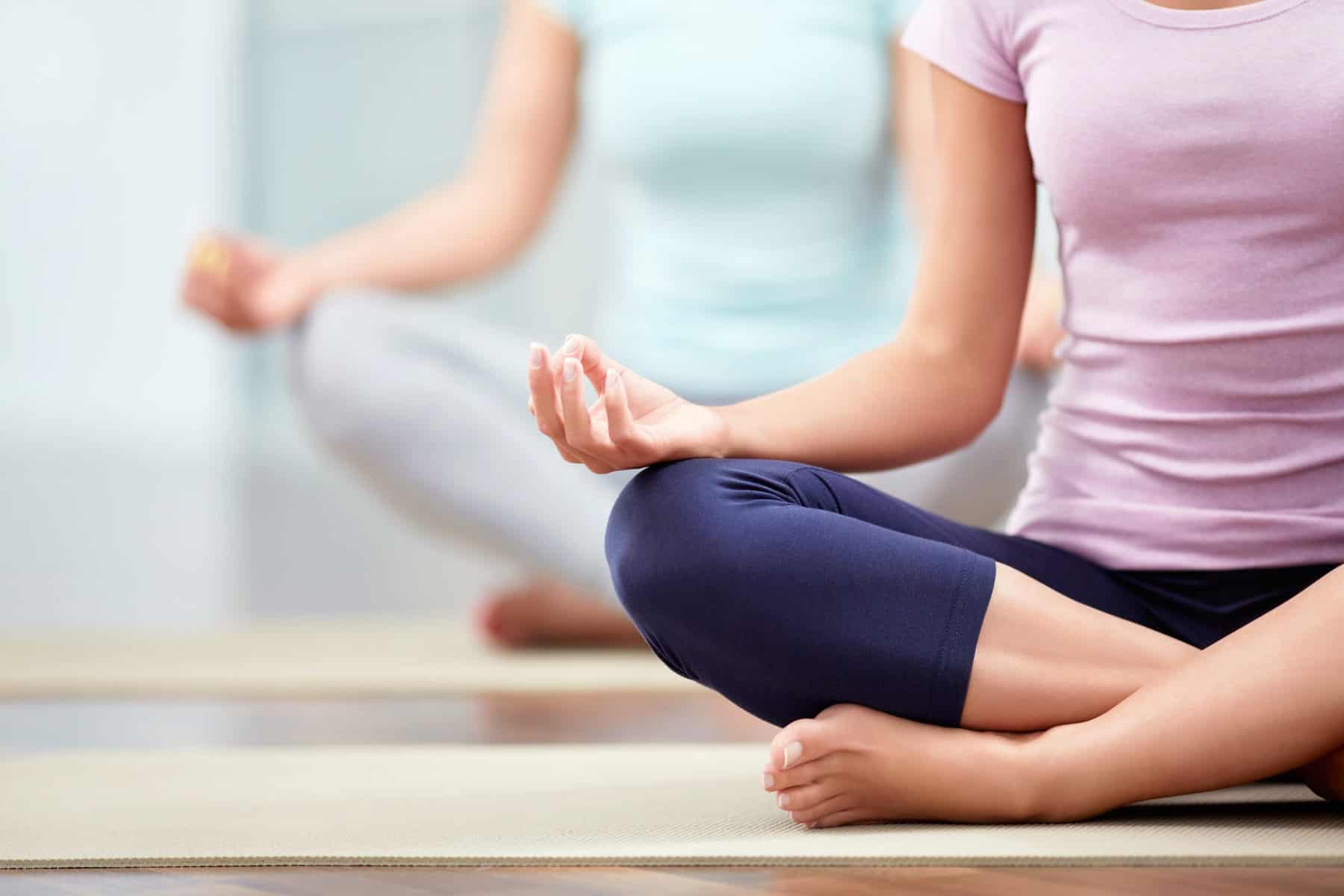 A cropped image of two woman seated in yoga mats in a cross legged position for easy pose with the palms on the knees and facing up for a yoga practice.