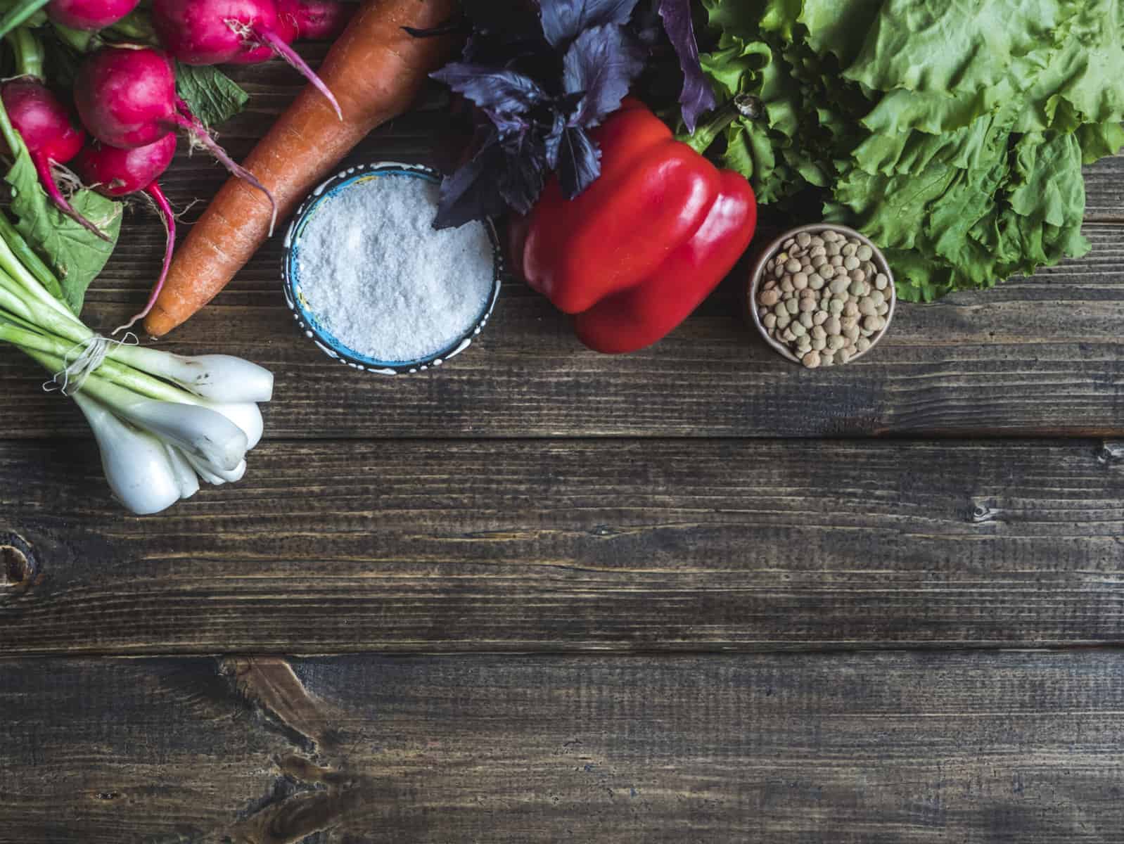 A wooden table with assorted frech vegetables and ingredients for healthy eating including a red pepper, fresh lettuce, scallions, and radishes.