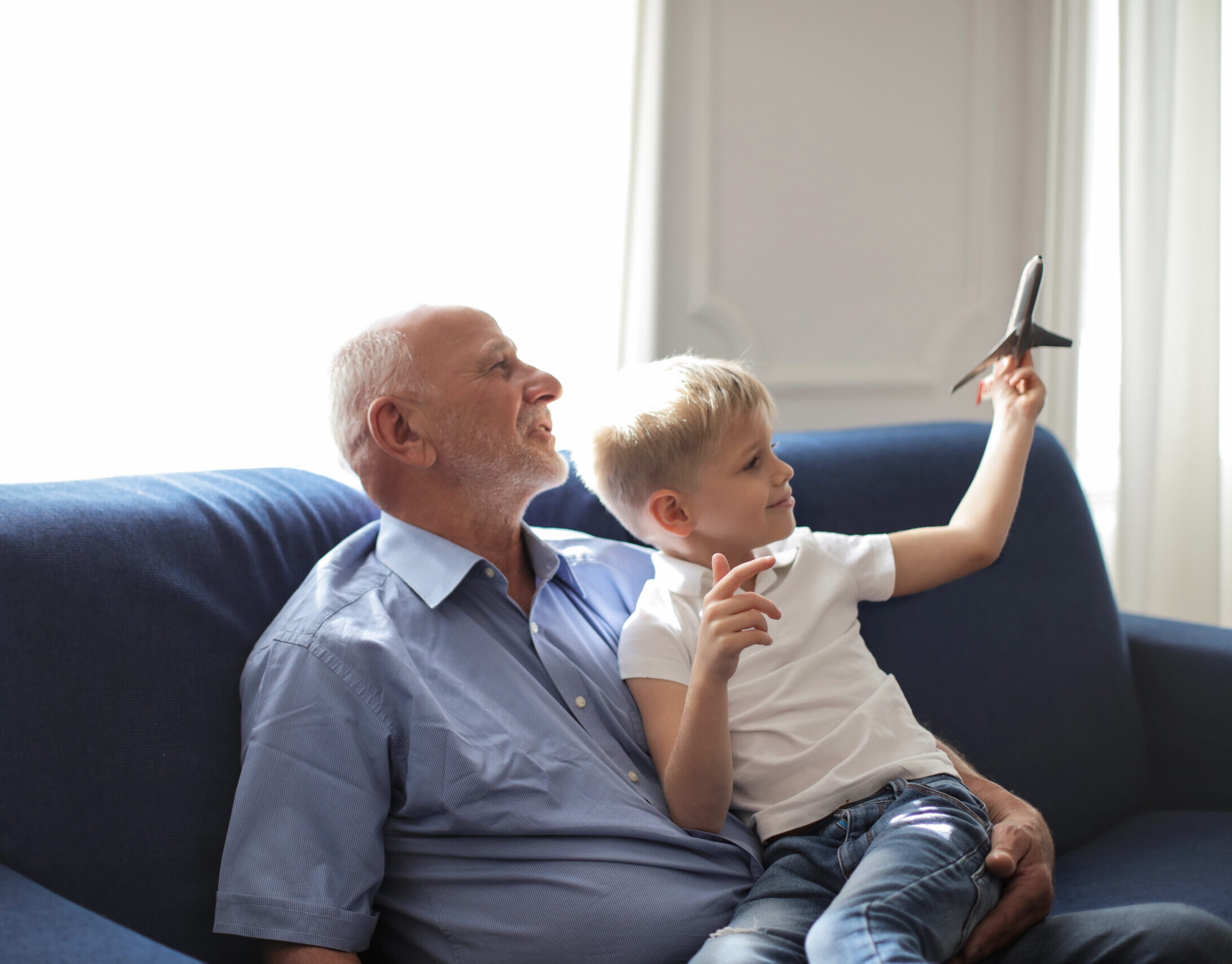 A grandparent and a grandchild sitting on a blue sofa near a window and playing with a toy airplane.