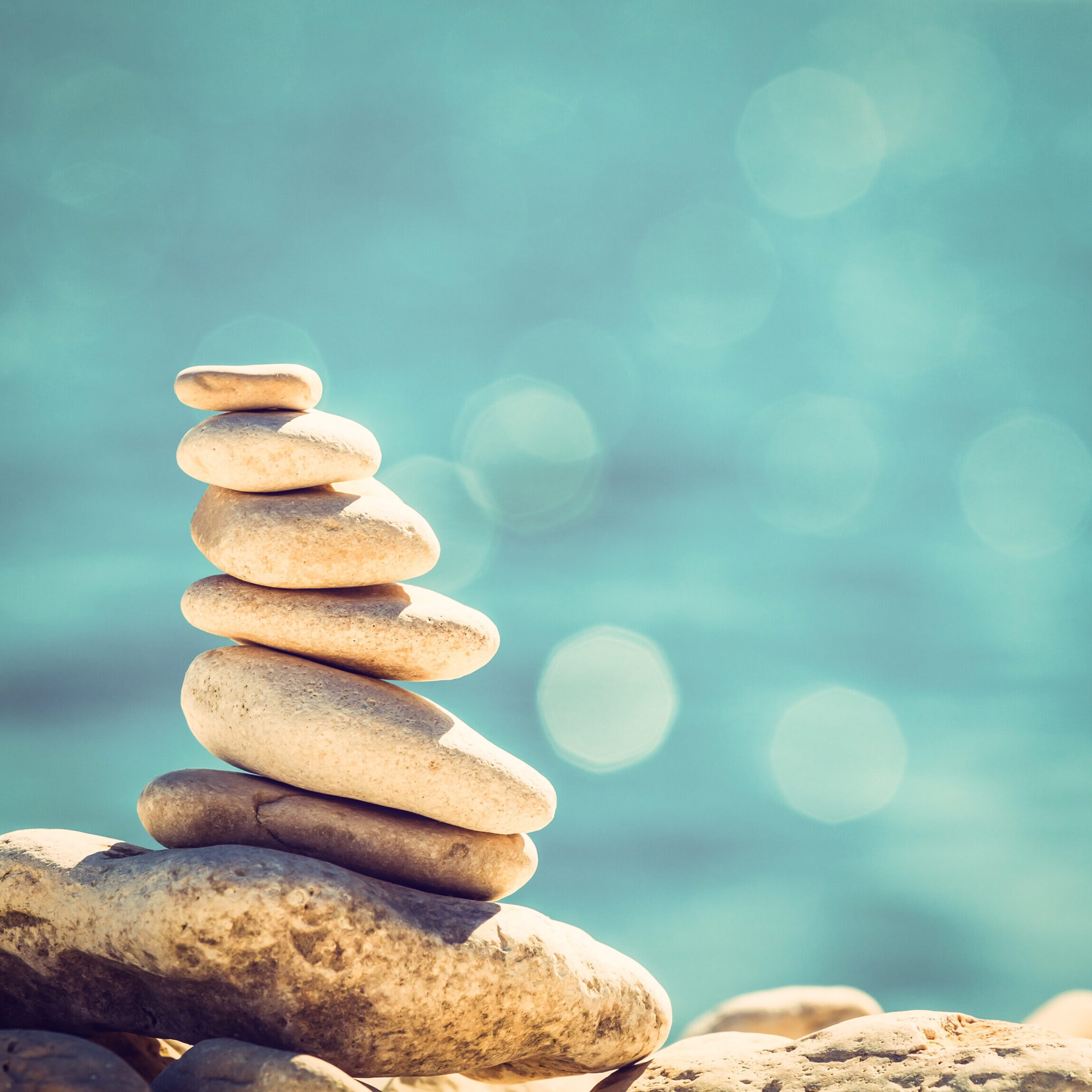 Stones balancing on a larger rock with a sparkling blue water in the background.