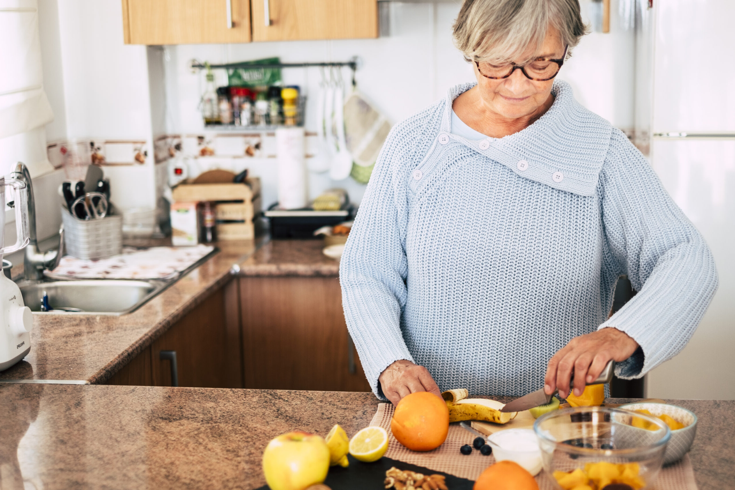 An older woman with glasses and a blue sweater standing at her kitchen counter while chopping fruit.