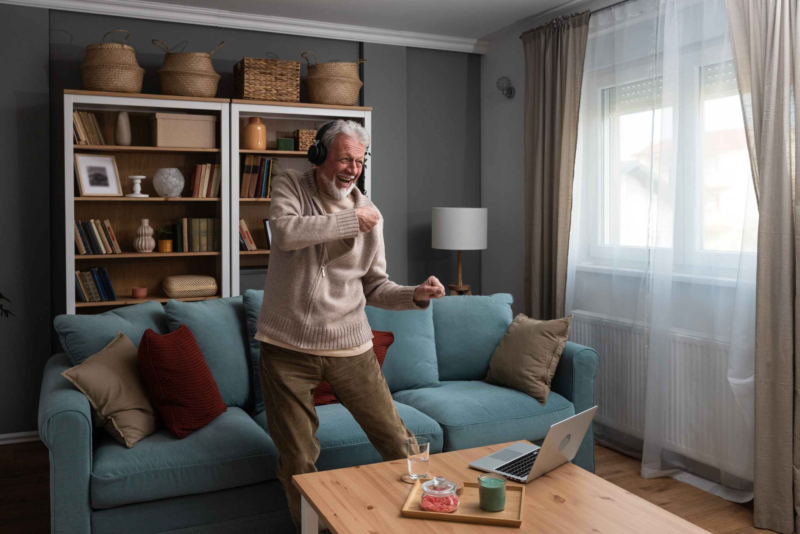 A happy smiling older man listening to music with wireless headphones standing up and dancing in a living room with a blue sofa and a large bookshelf.