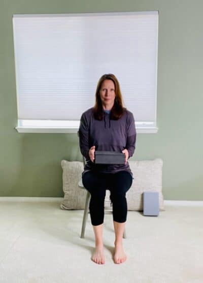 A woman seated in a chair near a window holding a yoga block in both hands above her thighs for a chair exercise.