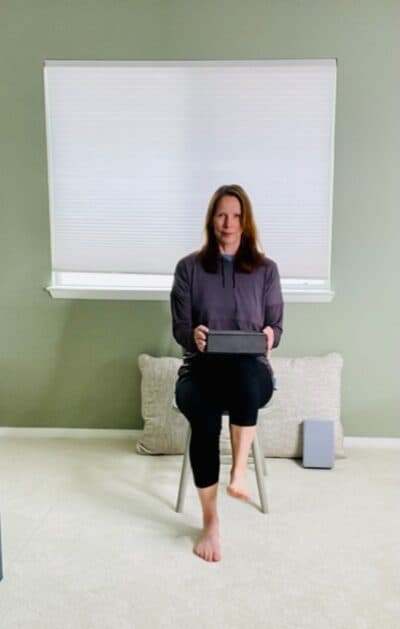 A woman seated in a chair near a window holding a yoga block above her thighs with one knee raised for a seated hip flexion exercise.