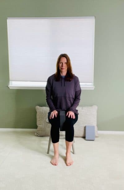 A woman seated in a chair near a window with a yoga block between her knees for a chair exercise.