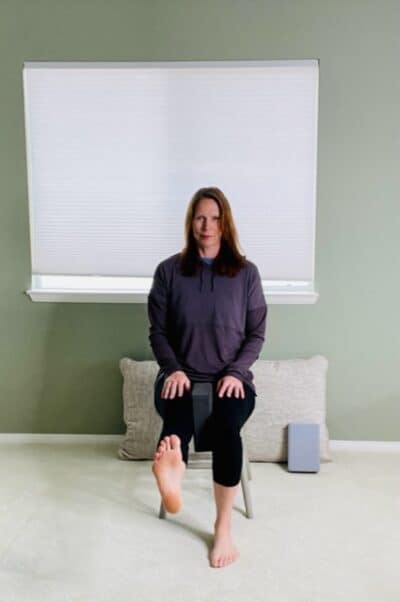A woman seated in a chair near a window with a yoga block between her knees and one knee extended for a chair exercise.