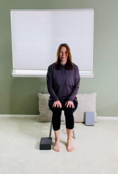 A woman seated in a chair near a window with a yoga block placed on the floor on the outside of one foot for a chair exercise.