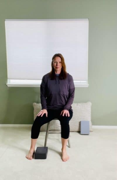 A woman seated in a chair near a window with a yoga block on the floor and one foot placed to the side for a chair exercise.