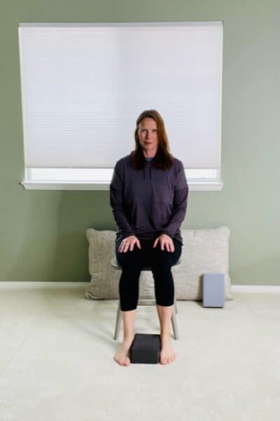 A woman seated in a chair near a window with a yoga block on the floor placed between her feet for a chair exercise.