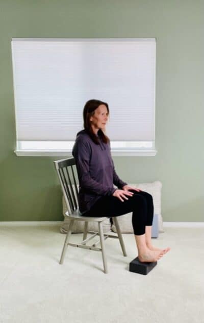 A woman seated in a chair near a window with her heels on a yoga block and her toes lifted up for an ankle dorsiflexion exercise.