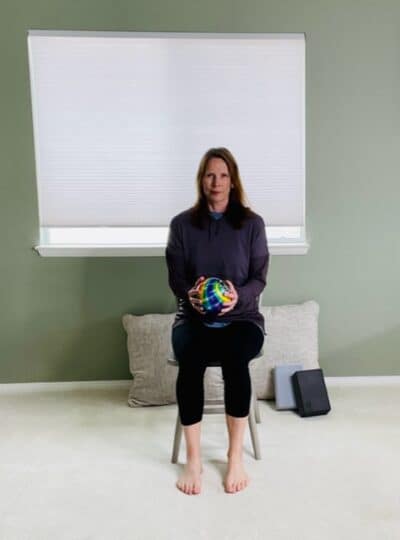 A woman seated on a chair in front of a window holding a ball with both hands in front of her body for a chair exercise.