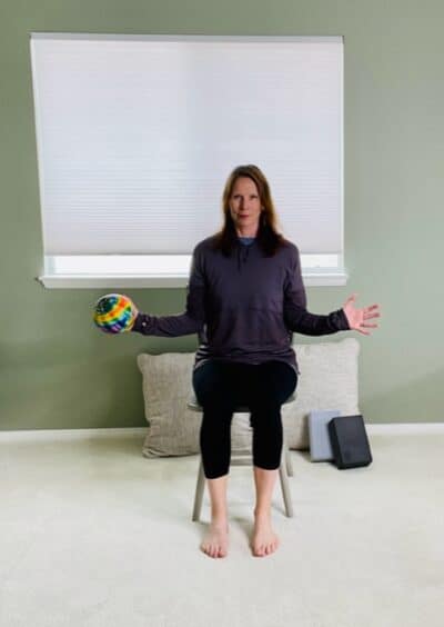 A woman seated in a chair near a window with both elbows bent and tucked at her sides while holding a ball on one hand for a shoulder external rotation exercise.