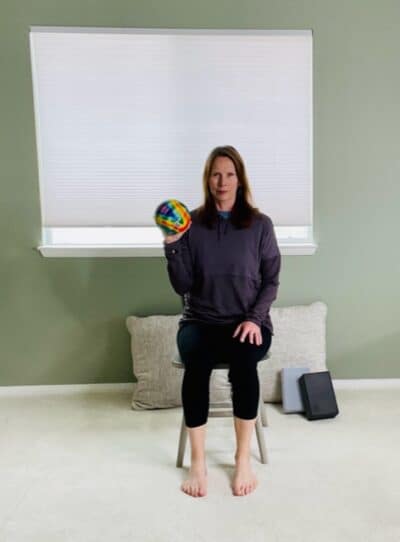 A woman seated in a chair near a window with one elbow bent and holding a ball in preparation for a chair exercise.