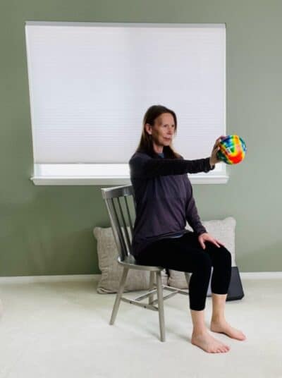 A woman seated in a chair near a window with one arm extended forward and holding a small ball for a chair exercise.