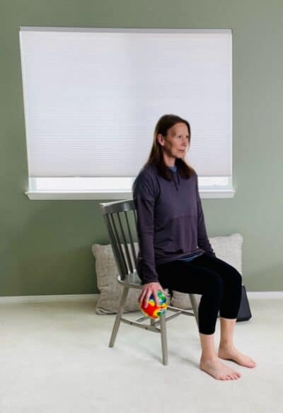 A woman seated in a chair near a window with her arms by her sides and holding a small ball in her right hand.