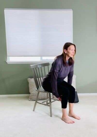 A woman seated in a chair near a window hinged forward at the hips while passing a small ball between her hands.