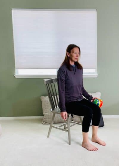 A woman seated in a chair near a window with her arms by her sides and holding a ball in her left hand for a chair exercise.