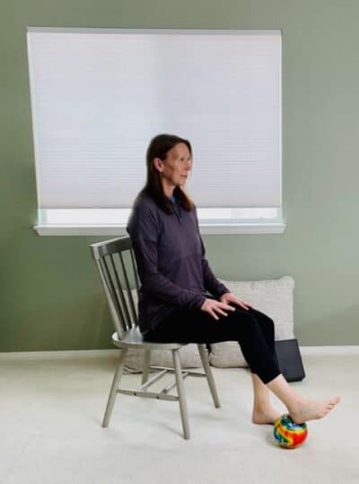 A woman seated in a chair near a window with one foot on a small ball under her heel for a seated chair exercise.