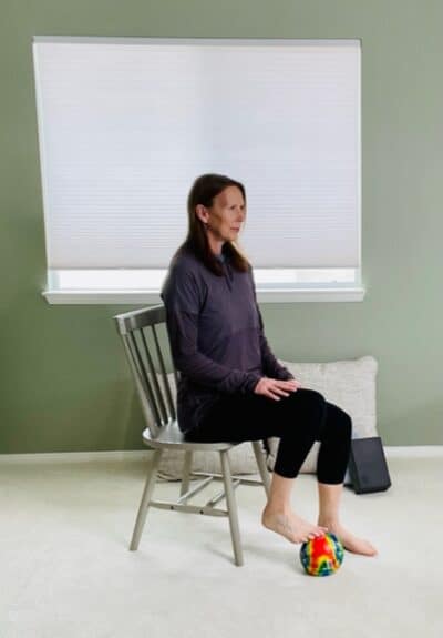 A woman seated in a chair near a window with one foot on a ball near her toes for a seated ball rolling exercise.