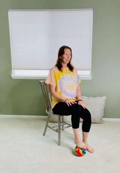 A woman seated in a chair near a window with one foot on top of a ball on the floor in preparation to a hamstring press exercise.