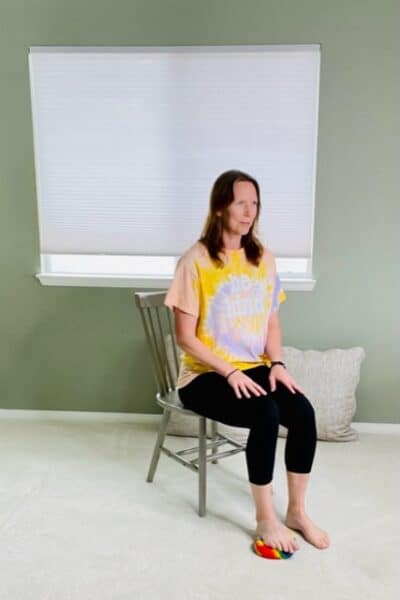 A woman seated in a chair near a window with a small ball under one foot as the presses down for a hamstring press exericse.