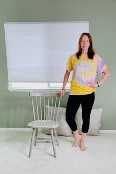 A woman standing next to a chair with one hand on the back of the chair, the other hand on the outside hip, and the inside leg externally rotated and raised up on the toes for a chair yoga tree pose variation.