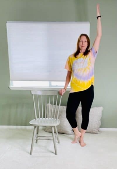 A woman standing next to a chair with one hand on the back of the chair for support, the other hand raised above her head, and the foot closest to the chair externally rotated and up on the toes for a chair yoga tree pose variation.