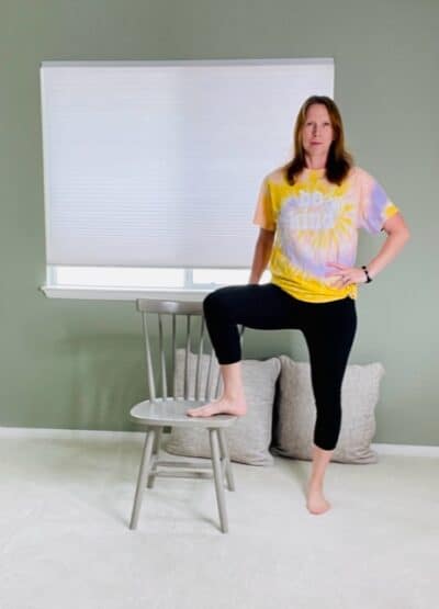 A woman standing next to a chair with one foot on the seat, one hand on the chair for support, and the other hand on her hip for a chair yoga tree pose variation.