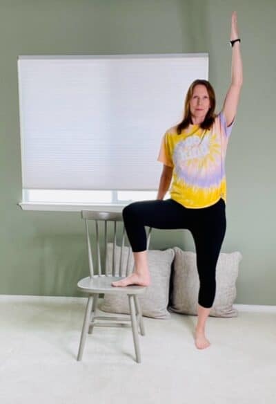 A woman standing next to a chair with one hand holding the back for support, one foot placed onto the seat of the chair, and the other hand raised up above the head for a chair yoga tree pose variation.