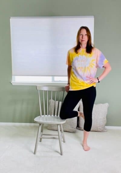 A woman standing next to a chair with one hand on the back of the chair, one knee resting on the seat of the chair, and the other hand on the hip for a chair yoga tree pose variation.