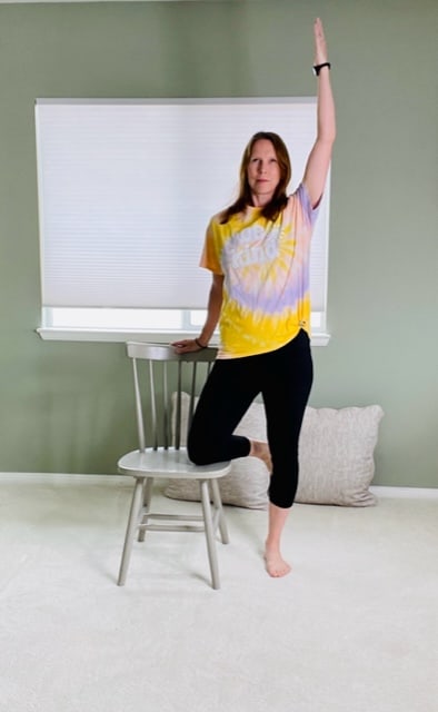 A woman standing next to a chair with one hand on the back of the chair, the inside knee resting on the seat of the chair, and the other arm raised overhead for a chair yoga tree pose variation.