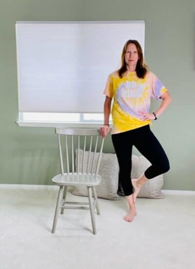 A woman standing next to a chair with one hand holding for support, the other hand on her hip and the foot of the outside leg resting on the calf on the standing leg for a chair yoga tree pose variation.