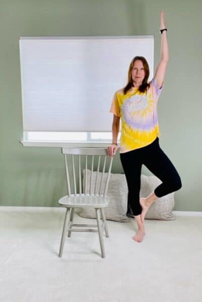 A woman standing next to a chair with one hand on the back of the chair, the other arm raised over head, and the outside foot resting on the calf of the standing leg for a chair yoga tree pose variation.