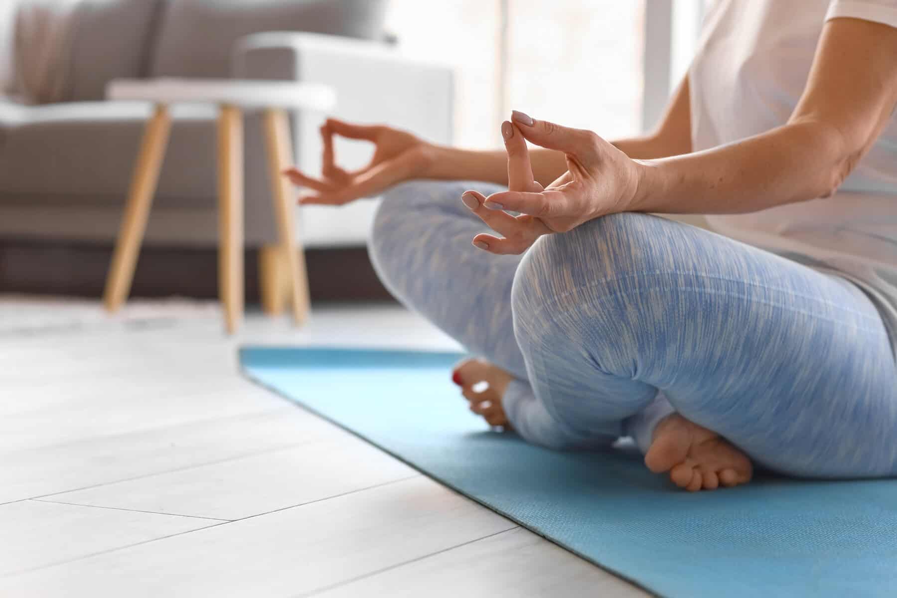 A cropped image on an older women seated on a blue yoga mat in a cross legged position with her hand on her knees and her thumbs touching her middle finger for a meditation pose.