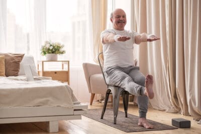 A senior man seated in a chair practicing a chair yoga pose in preparation for bird dog chair yoga pose.