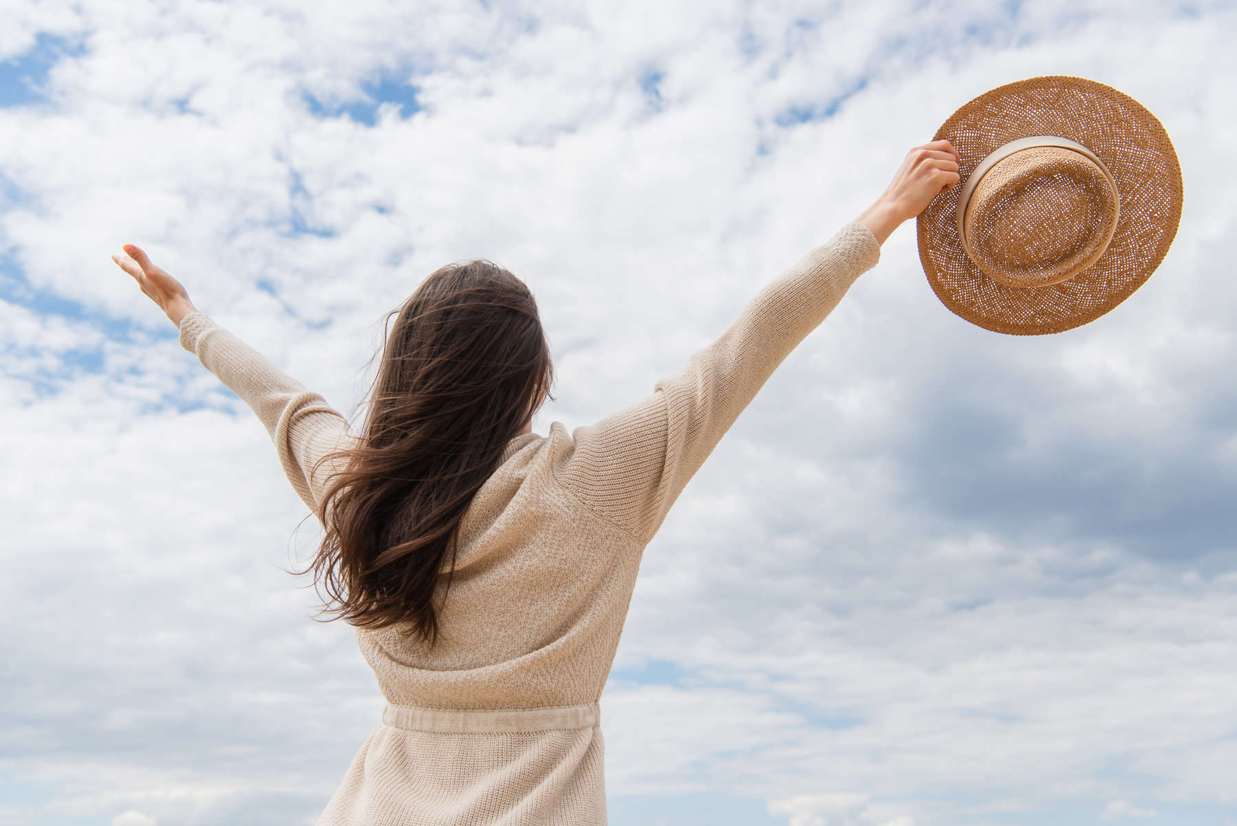 A young woman facing away with her hands stretched towards a cloud filled sky holding a hat in one hand to represent a positive attitude.