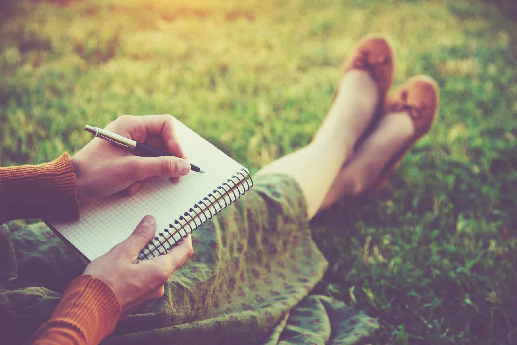 A cropped images of a woman sitting in the grass with a notebook and pen for journaling and goal setting and planning.