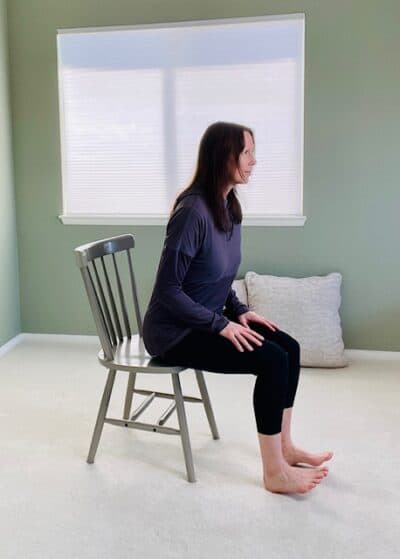 A women seated in a chair near a window practicing chair pose, hinged forward at the hips with one foot lifted up onto the heel.