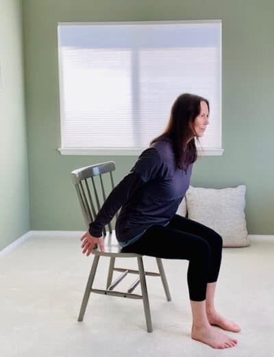 A women seated in a chair near a window practicing chair yoga chair pose with airplane arms, hinging forward at the hips with both arms extended slightly behind the body.