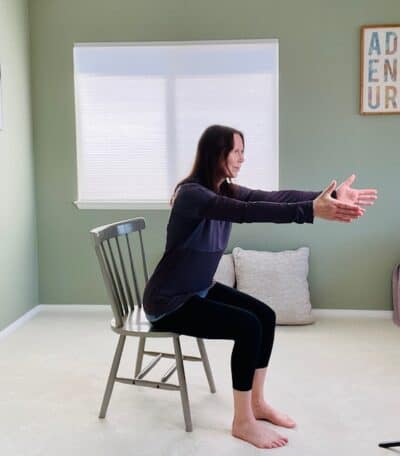 A woman seated in a chair near a window practicing chair yoga chair pose, hinging forward at the hips with her arms raised in front of her body.