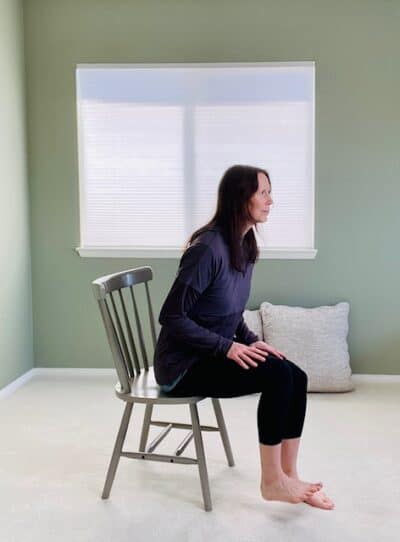 A woman seated in a chair near a window practicing a one Legged chair pose variation with one foot lifted up off the ground.