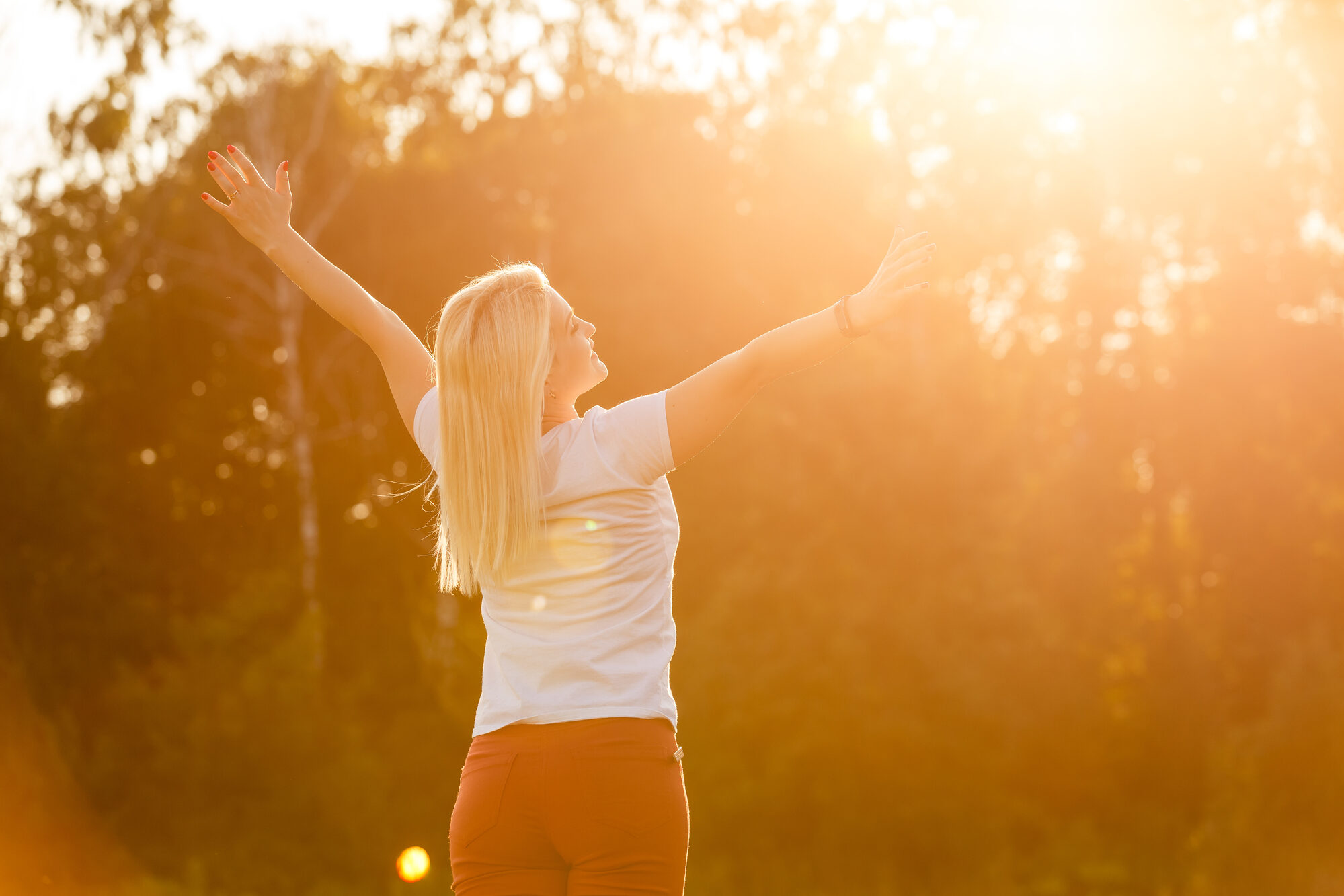 A woman in sunlit trees with her arms raised up and out to the side in an energetic pose.