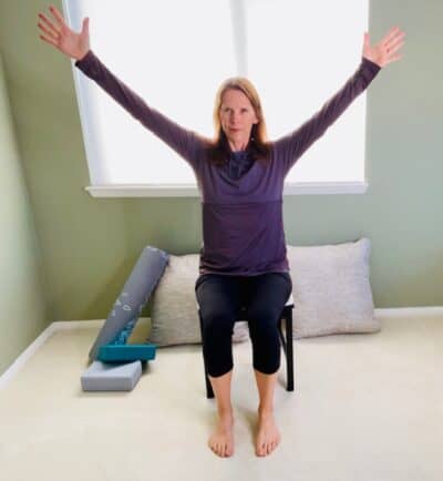 A woman seated in a chair near a window practicing the chair yoga pose five pointed star with both arms raised in a V shape.