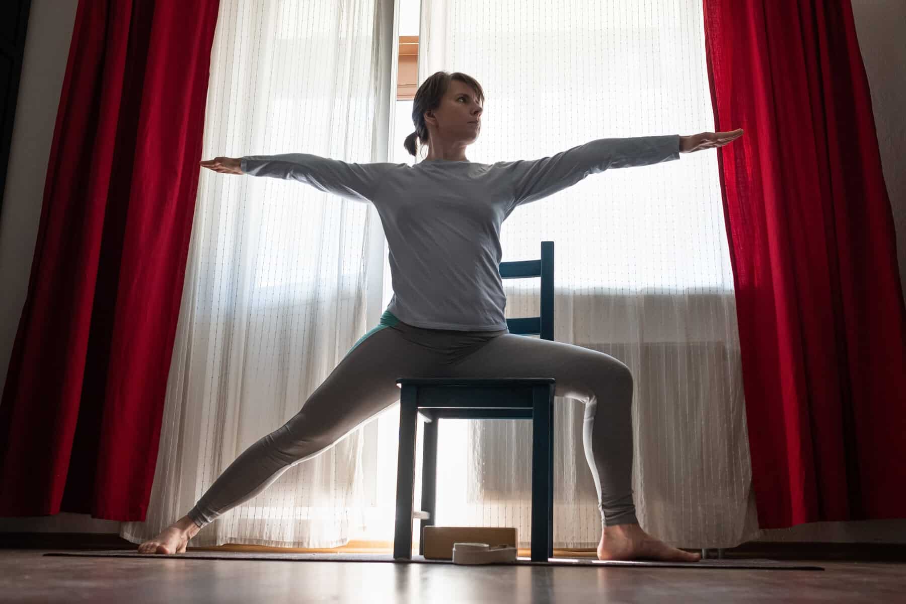 A woman practicing Warrior II pose in a chair in front of a sun lit window with both arms extended and one knee straight and the other bent.