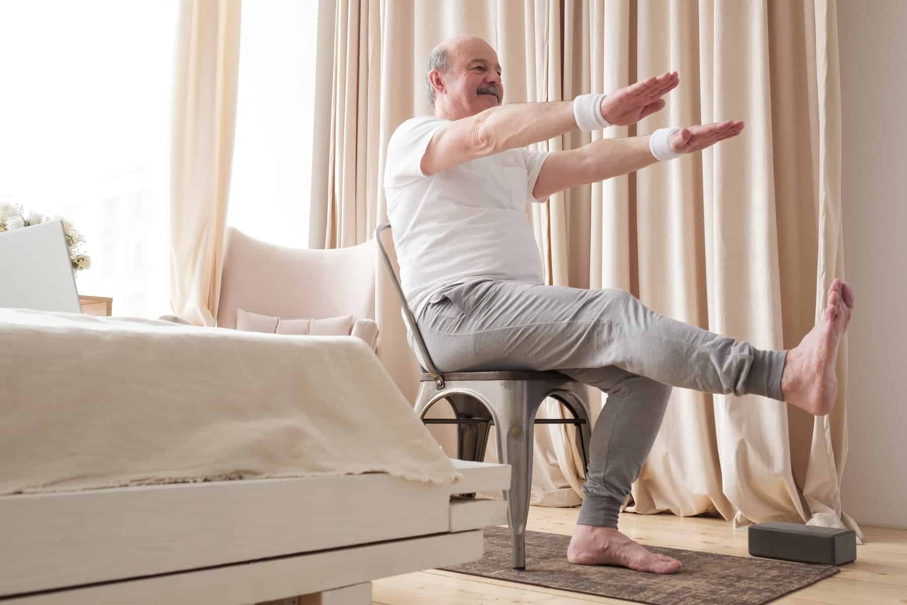 An older man seated in a chair near a window with a curtain with both arms raised in front of his body and one knee extended to practice chair yoga poses.