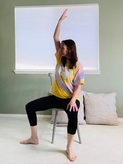 A woman seated in a chair near a window with one leg turned out the the side and the arm raised up to stretch the side for a Reverse Warrior Chair yoga pose.