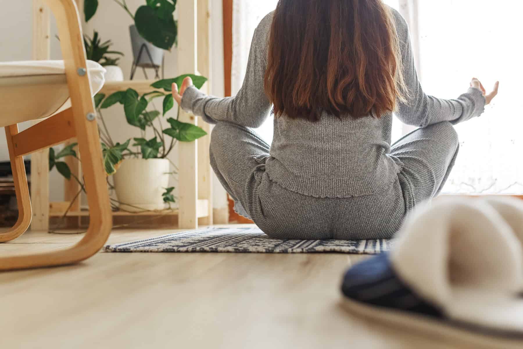 A women seated in yoga pose, easy pose, near a window next to a potted plant for meditation and mindfulness breathing practice.