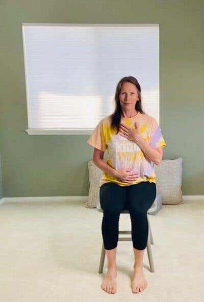 A woman seated in a chair with one hand on her belly and one hand on her chest for a yoga breathing exercise.