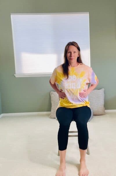 A woman seated in a chair near a window with her hands placed on her outer ribs on each side for a lateral breathing exercise.