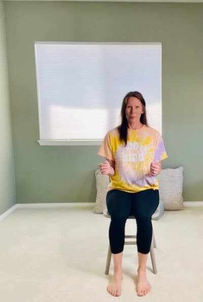 A woman seated in a chair near a window with her elbows bent at her low ribs for a chair yoga breathing exercise.